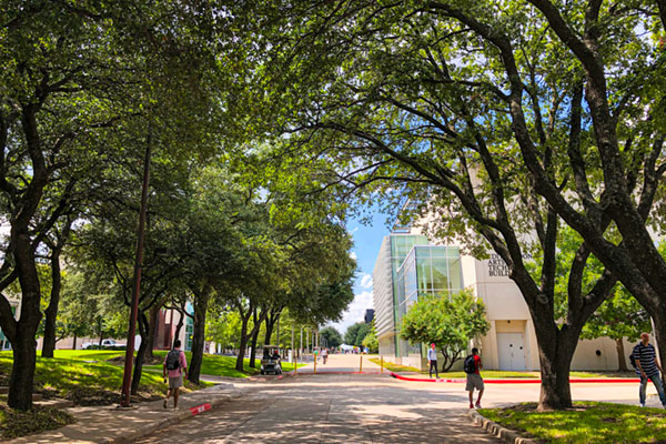 UT Dallas students walking under a canopy of trees near the JSOM and ATEC buildings